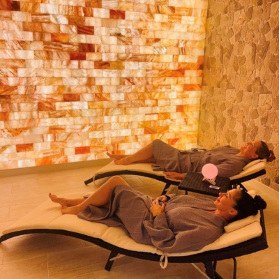 Two Woman In Lounge Chairs In A Salt Room With A Himalayan Salt Brick Wall.