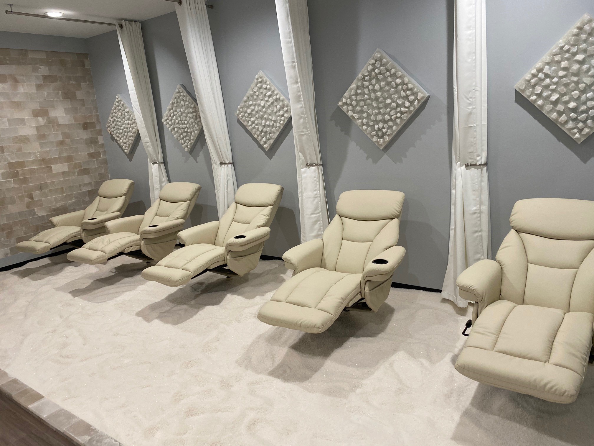 Five white cushioned reclining chairs on a salt-covered floor with a salt stone wall, four white held up separator curtains, and five diamond shaped slat stone decor on the walls.
