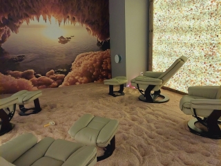 Multiple Tan Chairs And Ottomans In A Room With A Salt-Covered Floor, An Led Backlit Salt Wall On The Right And A Mural Of A Salt Cave On The Left.