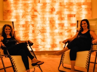 Two Smiling Women Sit In Lounge Chairs With A Himalayan Salt Brick Wall In The Background At The Salt Barre In Pittston, Pennsylvania