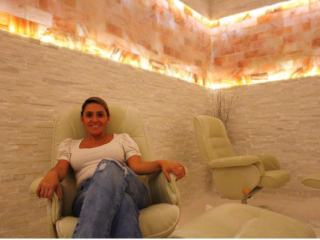 The Salt Nest. Woman In White Shirt And Jeans Sits In Chair And Poses For The Camera In Salt Room