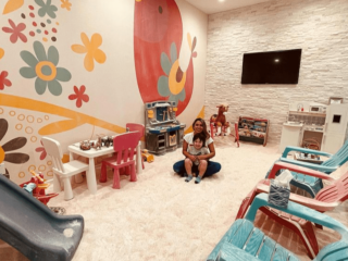 The Salt Nest. Woman and her son sit in the middle of the salt room. Around the room are adirondack chairs, toys, a slide, toy tool bench as a rocking horse. Mounted on the wall is a tv and a painted mural