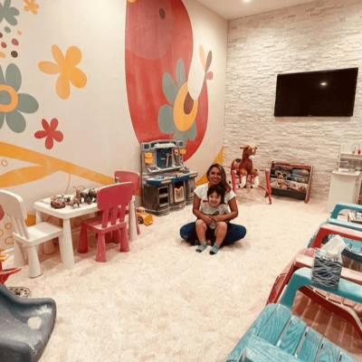 The Salt Nest. Woman and her son sit in the middle of the salt room. Around the room are adirondack chairs, toys, a slide, toy tool bench as a rocking horse. Mounted on the wall is a tv and a painted mural