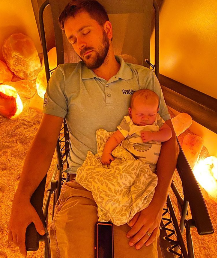 Eating Elevated. Man sitting in reclined chain with baby in his lap while in a lit salt room