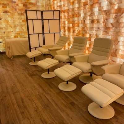 A Salt Room With Four Recliner Chairs, A Massage Table, And A Himalayan Salt Brick Wall.