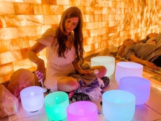 Prana Salt Cave. Women Who Appears To Be Leading A Meditation Session Has 7 Colorful Bowls Around Her. She Is Seen Knocking On A Bowl That Is Meant To Relax Those In The Session Who Can Be Seen Laying Down All The Way To The Right Of The Image,