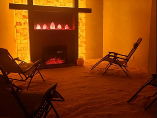 Radiant Well-Being. Four Lounge Chairs In Salt Room Facing A Fireplace. Above The Fireplace Are 5 Salt Rocks Which Are Glowing.