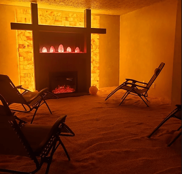 Radiant Well-Being. Four lounge chairs in salt room facing a fireplace. Above the fireplace are 5 salt rocks which are glowing.