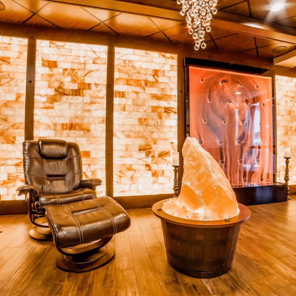 The Healing Den Of Salem. Cushioned Chair With Ottoman Next To Large Salt Rock Center Piece. Behind Them Are Salt Tiled Walls As Well As What Appears To Be A Glass Structure On The Back Wall With Salt Flowing Through It