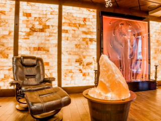 The Healing Den Of Salem. Cushioned Chair With Ottoman Next To Large Salt Rock Center Piece. Behind Them Are Salt Tiled Walls As Well As What Appears To Be A Glass Structure On The Back Wall With Salt Flowing Through It