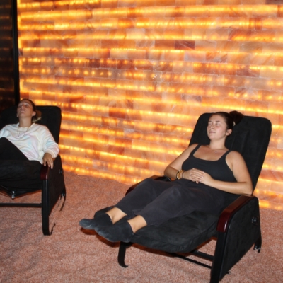 Bella Vita Cbd And Wellness. Two Women Laying Back In Lounge Chairs That Are In A Salt Room With Salt-Covered Floors And A Led Backlit Salt Brick Wall.
