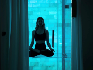 Breathe Meditation &Amp; Wellness. Woman Sitting Criss-Crossed In Glass Salt Chamber With A Bright Blue Ambient Light Glowing.