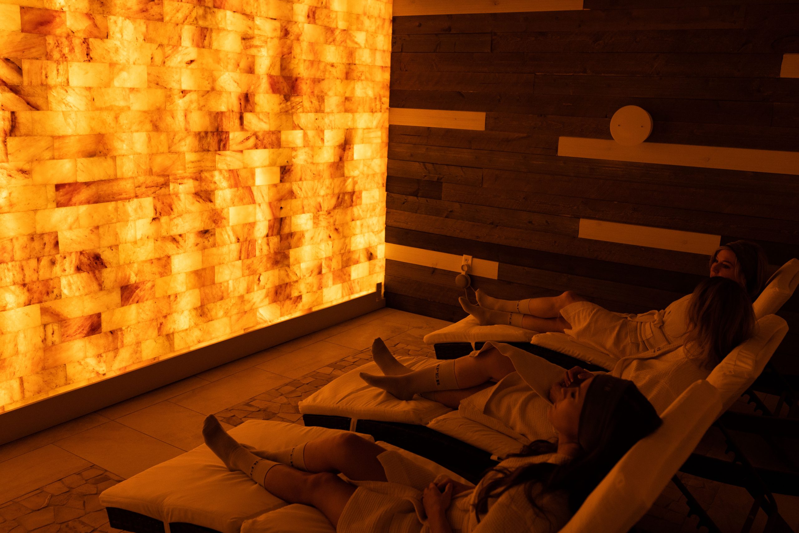 Three women relaxing on white chaises in front of an orange backlit salt wall at the SHarper the Medical Spa in Fishers, Indiana