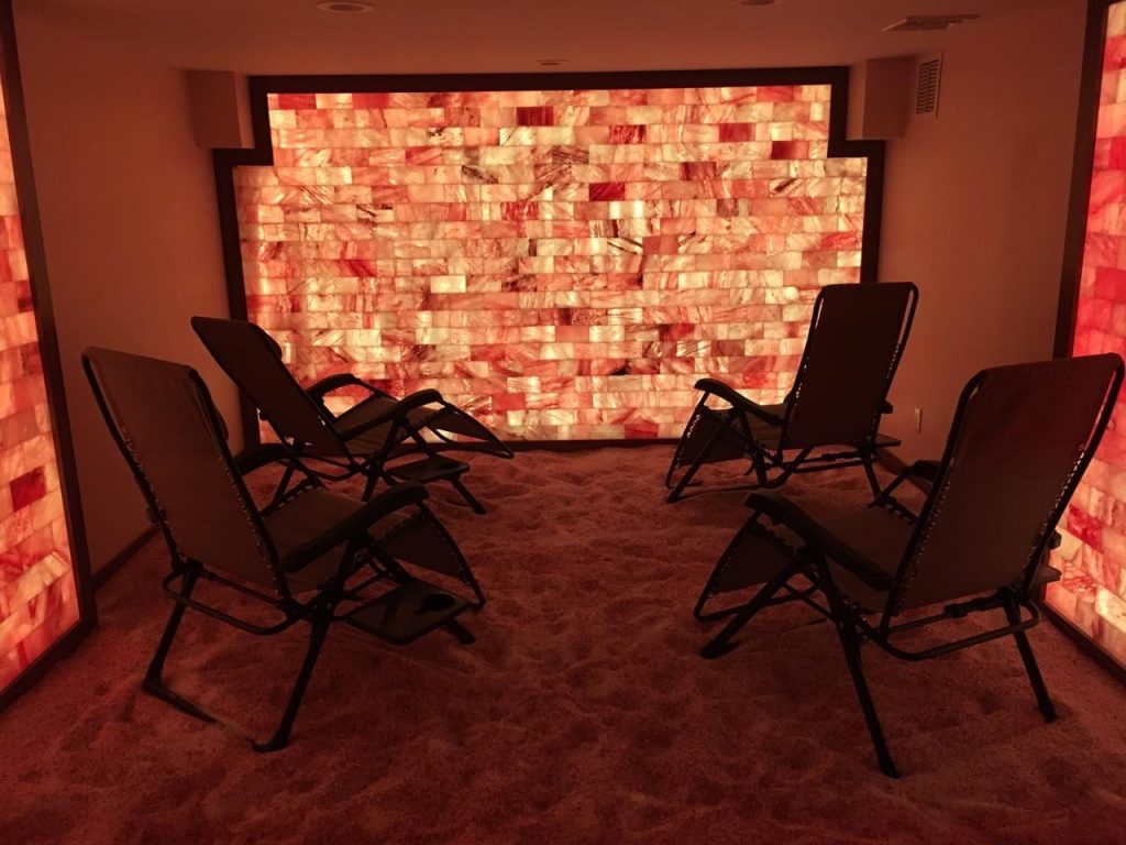 The Salt Therapy Room. Four Lounge Chairs Sitting On Top Of The Salt Covered Floor, All Face Towards The Front Of The Room Where The Wall Is Covered In Red And White Salt Tiles.