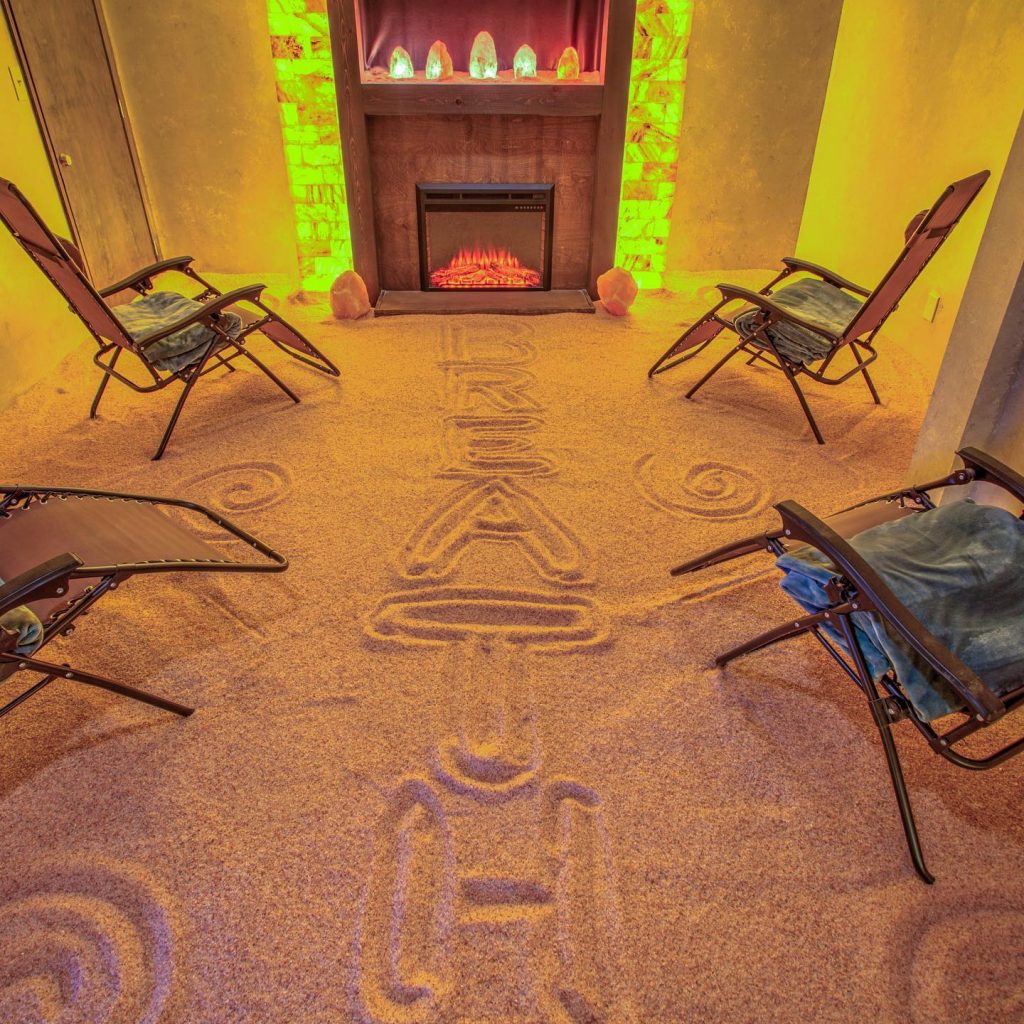 Radiant Well-Being. Four lounge chairs in salt room facing a fireplace. Above the fireplace are 5 salt rocks which are glowing. Through the middle of the room, the word "breathe" is written in the salt.