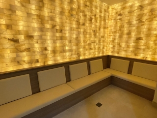 A Bench With White Cushions Against A Yellow Backlit Salt Stone Wall With White Flooring At The Renova Salon &Amp; Medspa In Carmel, Indiana