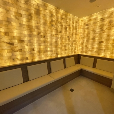 A bench with white cushions against a yellow backlit salt stone wall with white flooring at the Renova Salon & MedSpa in Carmel, Indiana