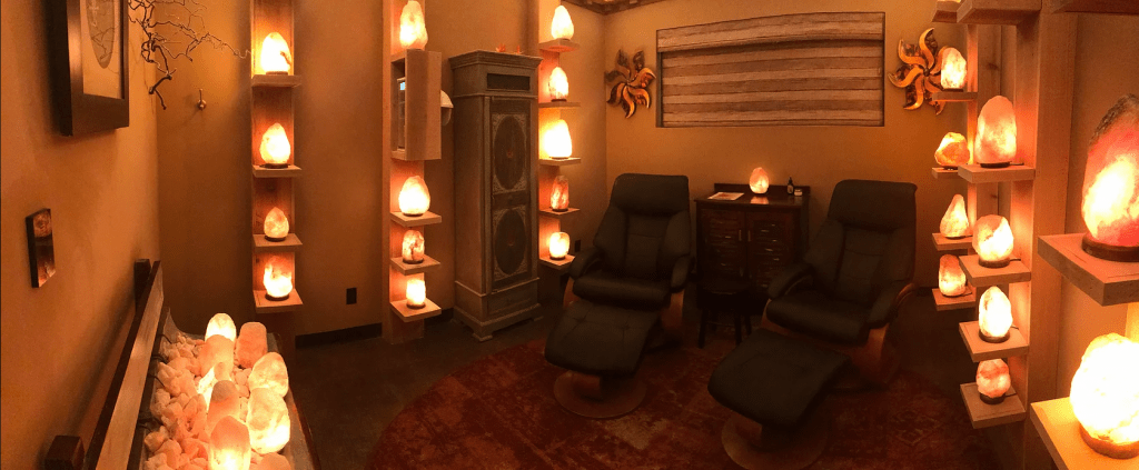 The Mandarin Spa At New Leaf. Dimly Lit Spa Room Has Two Chairs In It With Numerous, Glowing Salt Rocks On Shelves All Around The Room.