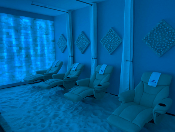 Four white cushioned reclining chairs on a salt-covered floor with a salt stone wall backlit by white lighting, four white held up separator curtains, and four diamond shaped slat stone decor on the walls.