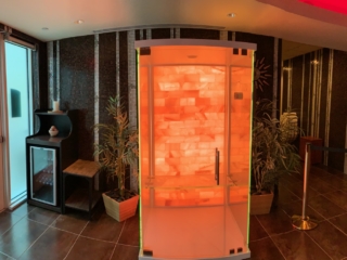 Salt And Sound Booth With A Salt Stone Wall Backlit With Orange Lighting On A Grey Tiled Floor With Two Plants Behind It Against A Tiled Wall.