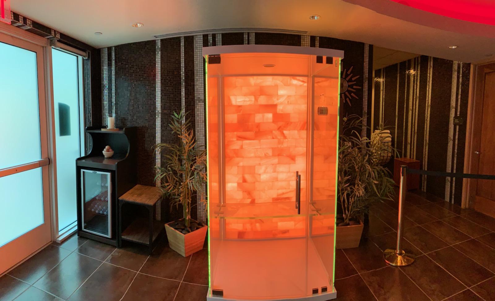 Salt and sound booth with a salt stone wall backlit with orange lighting on a grey tiled floor with two plants behind it against a tiled wall.