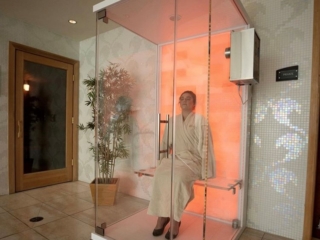 A Woman Relaxing In White Robe In A Glass Halotherapy Booth With A Salt Stone Wall At The Back Backlit With Orange Lighting Against A White Tile Wall.