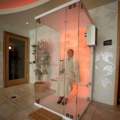 A woman relaxing in white robe in a glass halotherapy booth with a salt stone wall at the back backlit with orange lighting against a white tile wall.