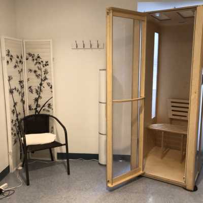 A wooden salt booth with a brown chair to the left against a folding panel with clothing hooks on the wall at the More Natural Health Center Ridgewood in New Jersey.