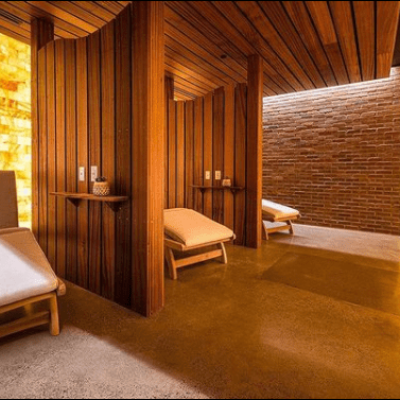 Salt Therapy Room With Three White Cushioned Chaises With Dark Wooden Dividers With An Orange Backlit Salt Wall Behind And In Front.
