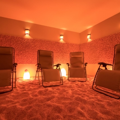 A Dimmed Lighted Room With Four Upright Chairs In A Salt Therapy Room Surrounded By A Pink Panel Of Salt Rocks With Pink Salt On The Ground At The Be Still &Amp; Breathe Salt Wellness Center.