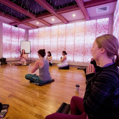 Five Women Sitting Crisscrossed In A Yoga Position In A Room With Purple And White Led Lit Salt Brick Walls And Panels At The Rise Yoga Spring Fest At Better Bodies Yoga.