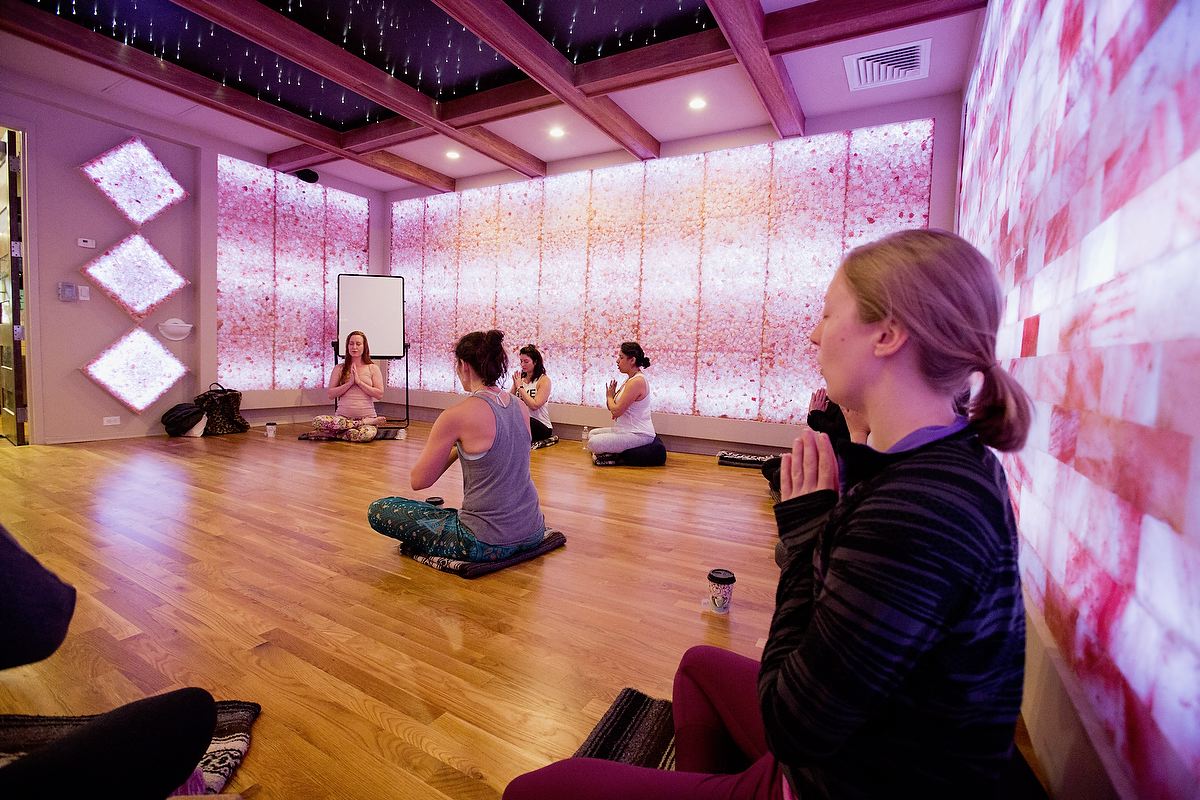 Five Women Sitting Crisscrossed In A Yoga Position In A Room With Purple And White Led Lit Salt Brick Walls And Panels At The Rise Yoga Spring Fest At Better Bodies Yoga.