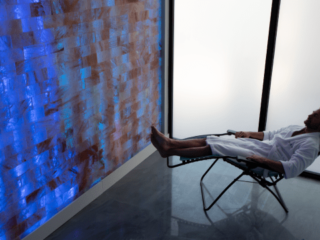 Man In White Robe Reclining In Chair In Front Of A Salt Brick Wall Backlit By Led Lights At Boca Cryo In Boca Raton, Florida