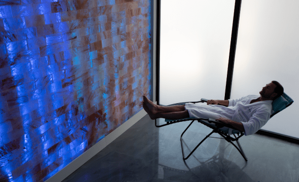 Man in white robe reclining in chair in front of a salt brick wall backlit by LED lights at Boca Cryo in Boca Raton, Florida