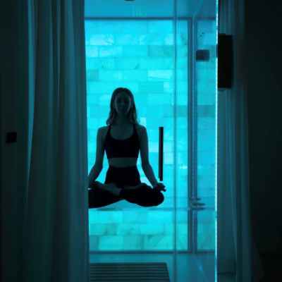 Woman sitting cross-legged in a glass salt and sound booth with a salt stone wall backlit with blue light.