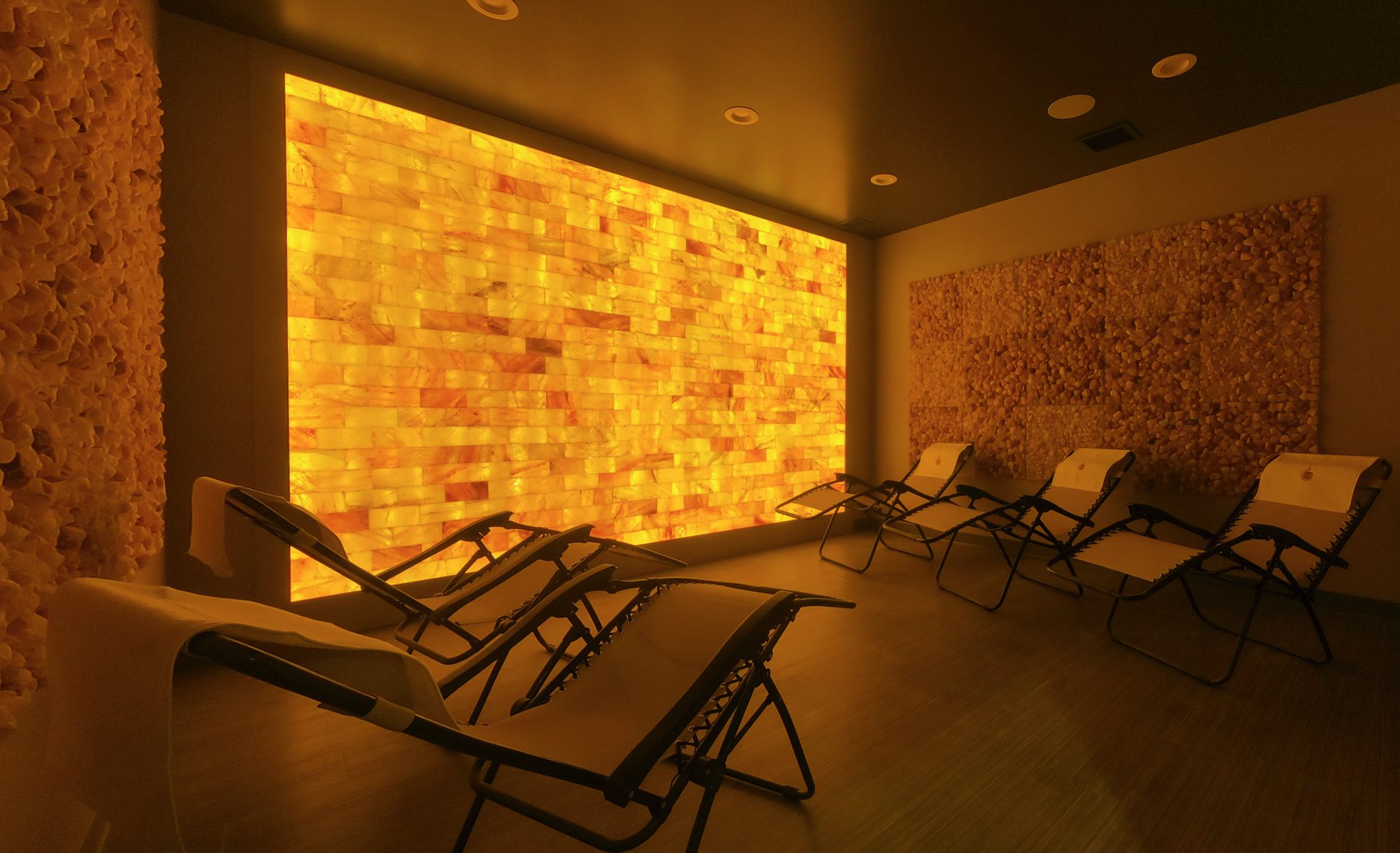 Five Reclining Chairs On A Wooden Floor In Front Of A Led Backlit Salt Panel Wall And Two Large Rectangular Salt Brick Wall Décor At The Breathe Salt Vault - Springfield, Missouri.