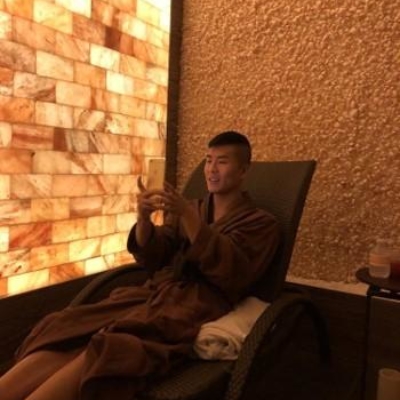 Man In Brown Robe Sitting On His Phone In Front Of A Himalayan Salt Stone Wall And A Paneled Backlit Orange Salt Wall.