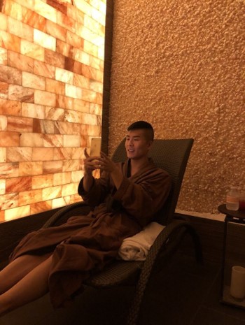 Man In A Brown Robe On His Phone Reclined In A Chair With A Salt Brick Wall Backlit By Led Lights.