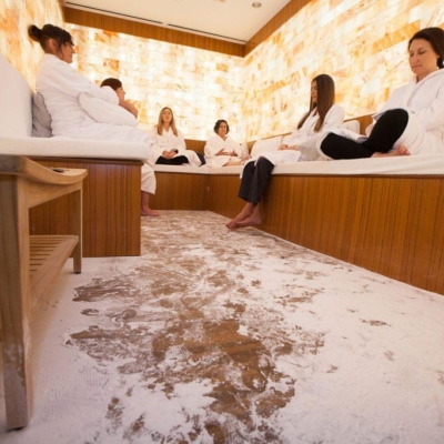 Six Women In White Robes Relaxing On A Booth With White Cushions And Pillows In A Room With White Salt-Covered Floors And A Backlit Salt Paneled Wall.
