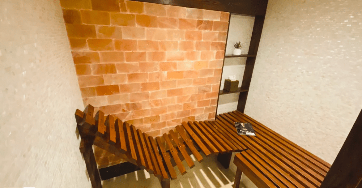 a light brown bench and lounge chair surrounded by white tiles and a Himalayan salt panel wall at Costa d'este Beach Resort & Spa - Vero Beach, Florida