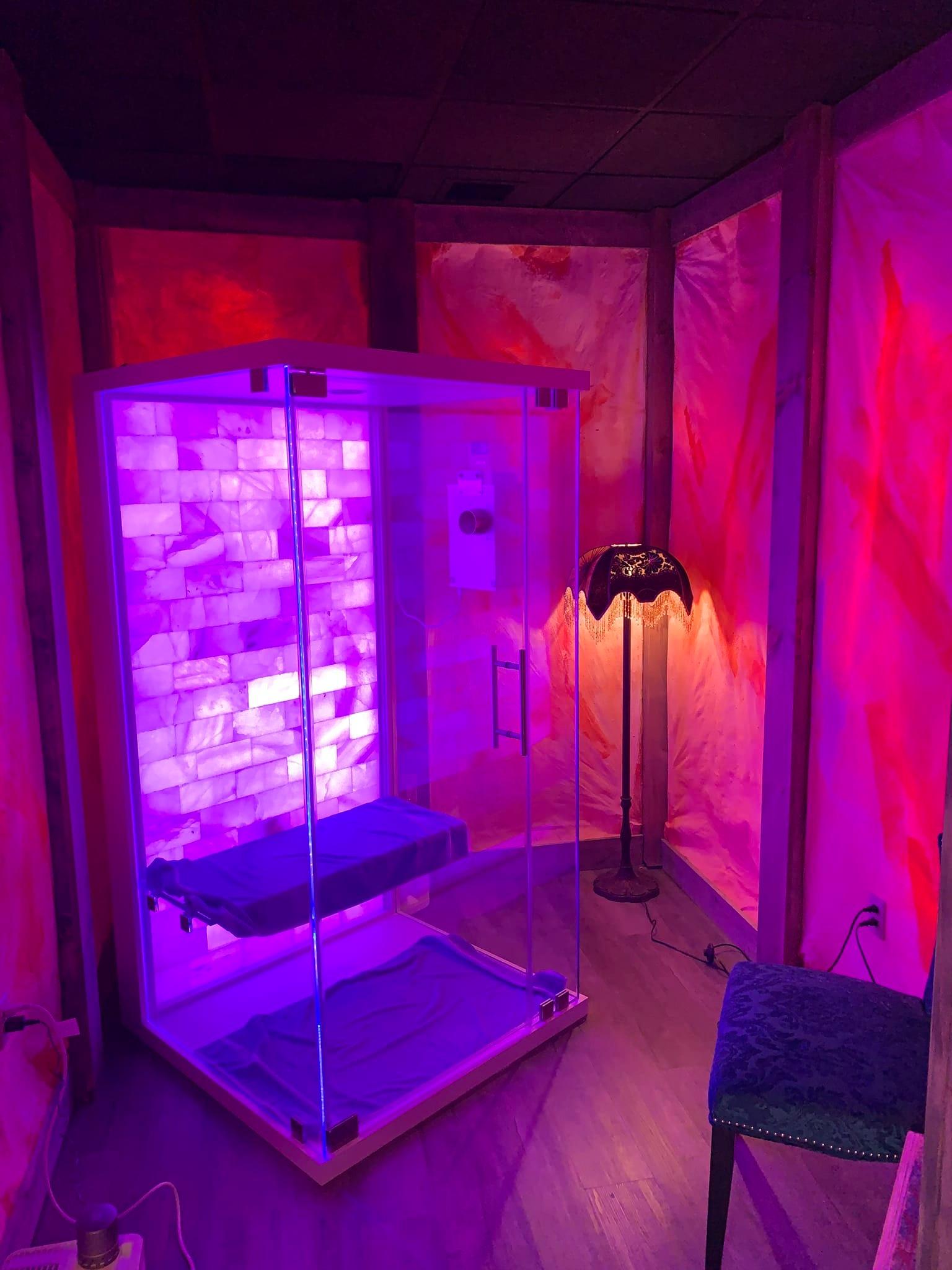 Glass halotherapy booth with a salt stone wall backlit with purple lighting in a paneled room backlit with purple and orange lighting at the Cryotherapy Plus Luxury Spa in Akron Ohio
