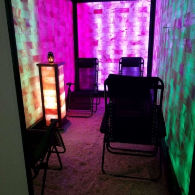 A Halotherapy Room With Four Reclining Chairs On A Salt-Covered Floor Surrounded By Paneled Salt Stone Walls Backlit With Green, Purple, White, And Orange Lighting.