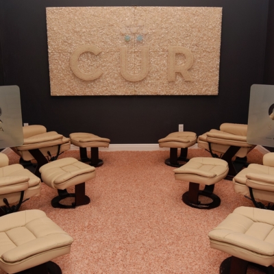 Six Tan Reclined Chairs All With A Foot Rest In A Dark Grey Room With The Cur Salt Spa Logo Personal Dividers And A Rectangular Salt Wall With The Cur Logo At The Cur Salt Spa Stuart Florida.