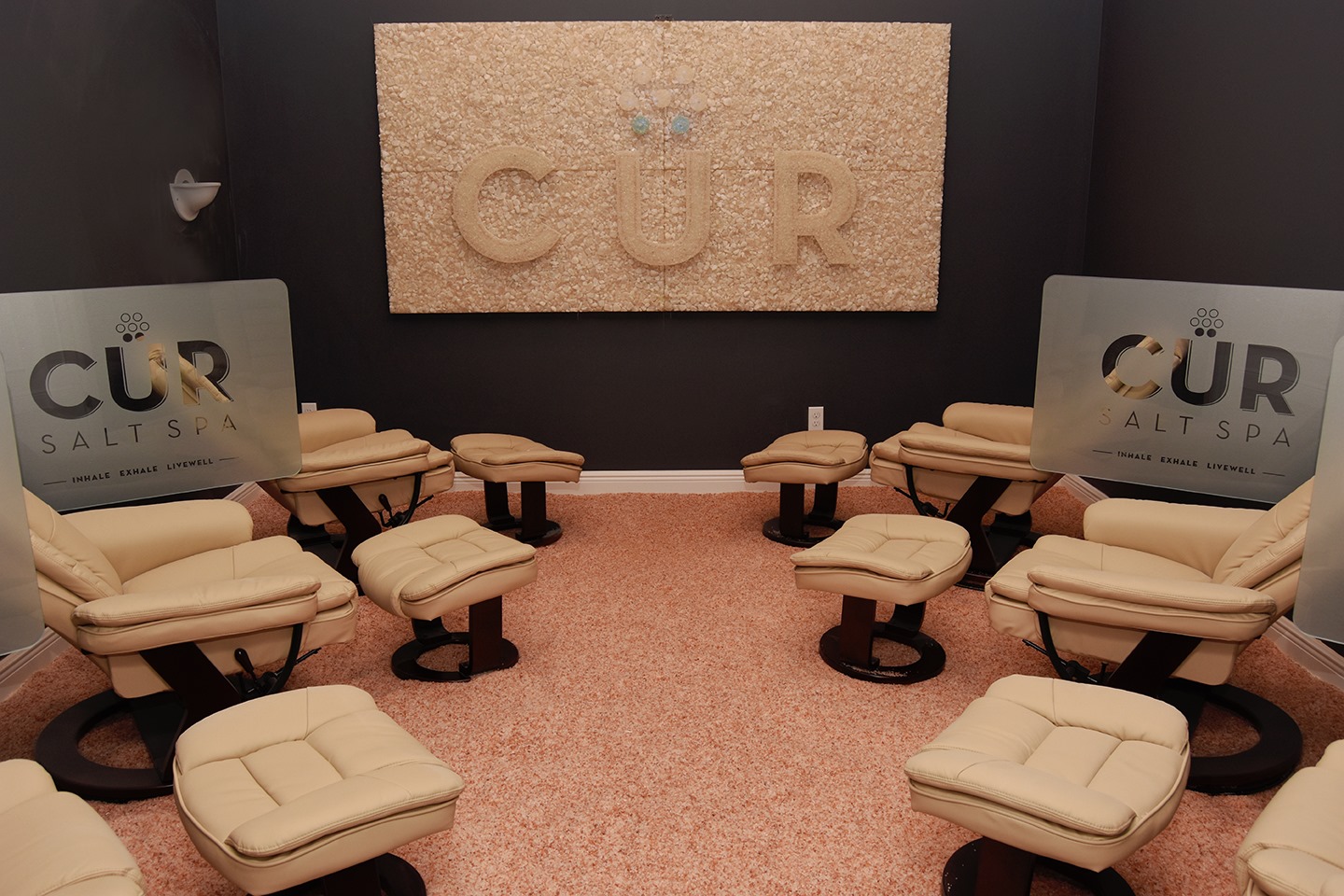 Four Cushioned Lounge Chairs With Dividers That Say &Quot;Cur Salt Spa Inhale Exhale Livewell&Quot; On A Salt-Covered Floor With A Large Rectangular Salt Brick Décor That Has &Quot;Cur&Quot; On It.