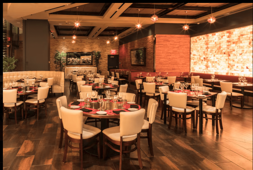 A Restaurant Dining Room With White Chairs And Tables With A Salt Brick Wall Backlit By Led Lighting.