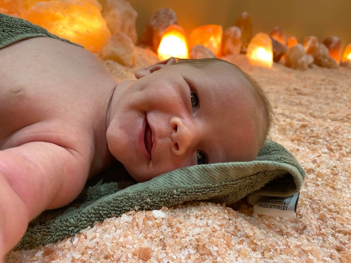 A smiling baby laying on a green towel on a salt covered floor with salt rock lamps in the background.