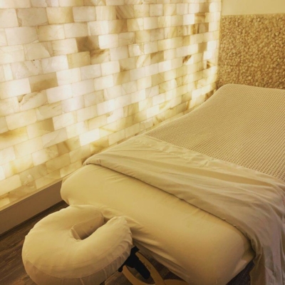 A White Cushioned Massage Table In Front Of A White Backlit Salt Stone Wall And Himalayan Salt Stones At Eleventh Element In Edwardsville, Pennsylvania