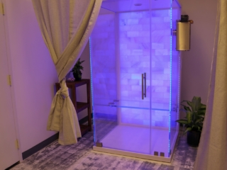 Glass Salt Therapy Booth With A White Salt Wall And Purple Led Lights On A White And Grey Carpet.