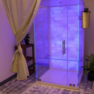 Glass salt therapy booth with a white salt wall and purple LED lights on a white and grey carpet.