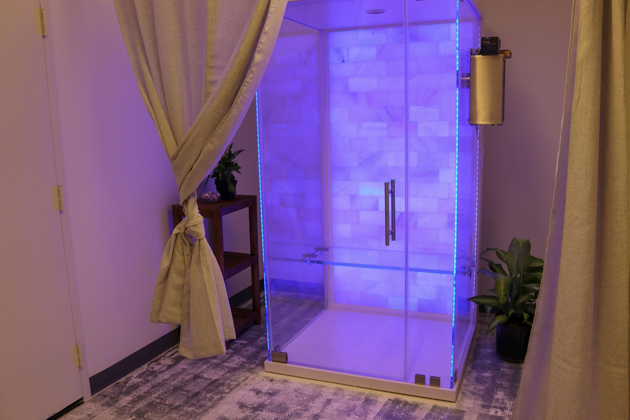 Glass salt therapy booth with a white salt wall and purple LED lights on a white and grey carpet.
